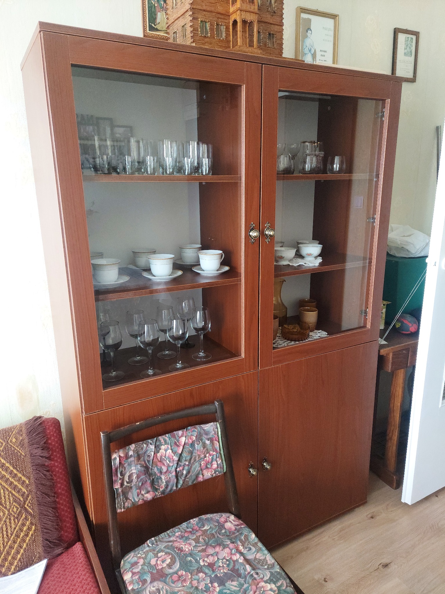 Dwudrzwiowa witryna z brązowego drewna, wypełniona szkłem, eksponująca zastawę stołową i dekoracje. Krzesło z kwiatowym wzorem stoi przed meblem.