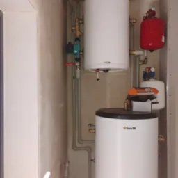 Dwa białe bojlery, czerwony zbiornik wyr&oacute;wnawczy i szare rury w kotłowni. Widoczne zawory i elementy instalacji. Na g&oacute;rnym bojlerze naklejka informacyjna.