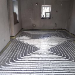 Michał Nieszporek - Białe rury ogrzewania podłogowego ułożone spiralnie na czarnej siatce w surowym wnętrzu z oknem. Żółta taśma brzegowa wzdłuż ścian.