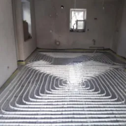 Białe rury ogrzewania podłogowego ułożone spiralnie na czarnej siatce w surowym wnętrzu z oknem. Żółta taśma brzegowa wzdłuż ścian.