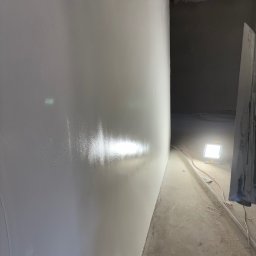 Oskar Polak Wykończenia Wnętrz - Gładka, jasna ściana po nałożeniu gładzi w nowym pomieszczeniu. Widoczne rury pod sufitem, lampa oświetlająca podłogę i narzędzia.