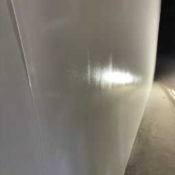 Oskar Polak Wykończenia Wnętrz - Świeżo pomalowana, gładka, jasnoszara ściana w rogu pomieszczenia, z widocznym połyskiem mokrej farby i nierównościami w strukturze. U dołu fragment szarej podłogi.