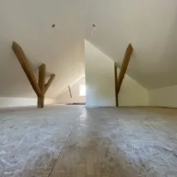 Poddasze z drewnianymi belkami i płytami OSB na podłodze, przygotowane do dalszych prac wykończeniowych. Białe ściany i sufit, widoczne punkty oświetleniowe.