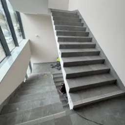 Wykończenie klatki schodowej z szarymi płytkami imitującymi kamień. Widoczni pracownicy podczas fugowania. Duże okno doświetla przestrzeń. Minimalistyczny styl.