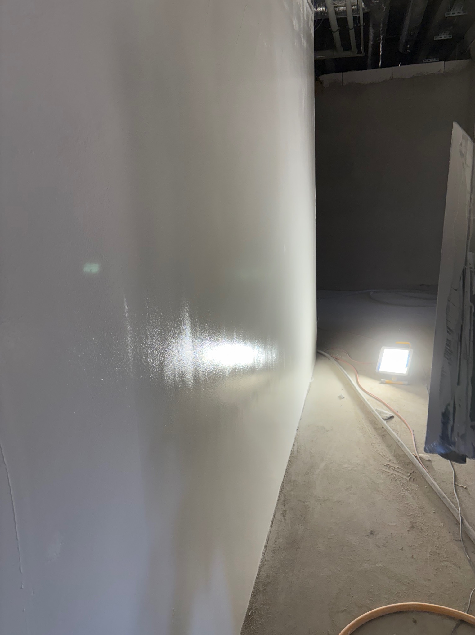 Gładka, jasna ściana po nałożeniu gładzi w nowym pomieszczeniu. Widoczne rury pod sufitem, lampa oświetlająca podłogę i narzędzia.