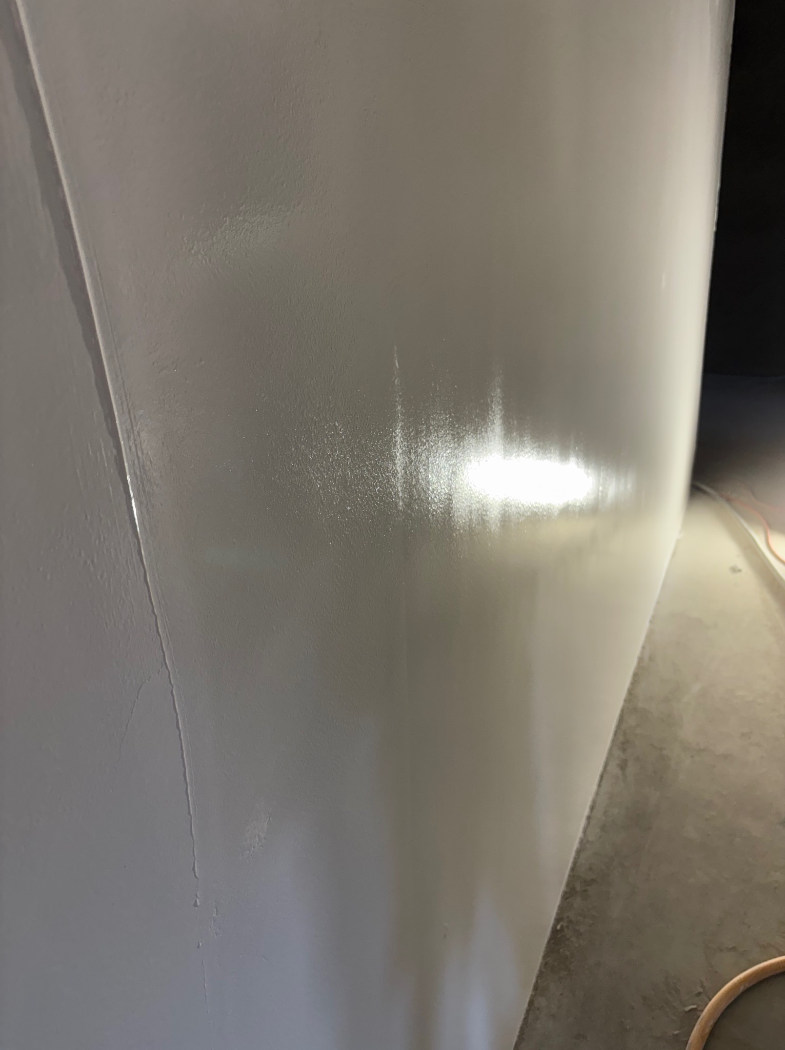 Świeżo pomalowana, gładka, jasnoszara ściana w rogu pomieszczenia, z widocznym połyskiem mokrej farby i nierównościami w strukturze. U dołu fragment szarej podłogi.