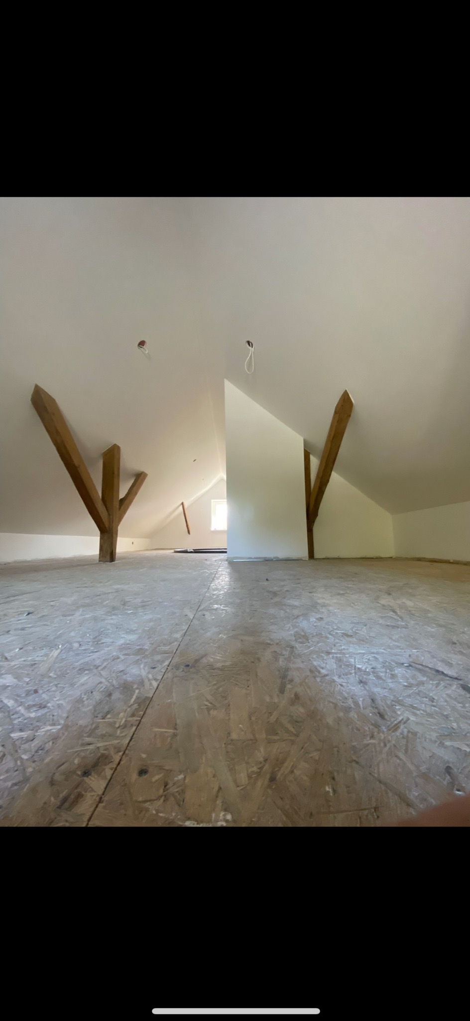 Poddasze z drewnianymi belkami i płytami OSB na podłodze, przygotowane do dalszych prac wykończeniowych. Białe ściany i sufit, widoczne punkty oświetleniowe.