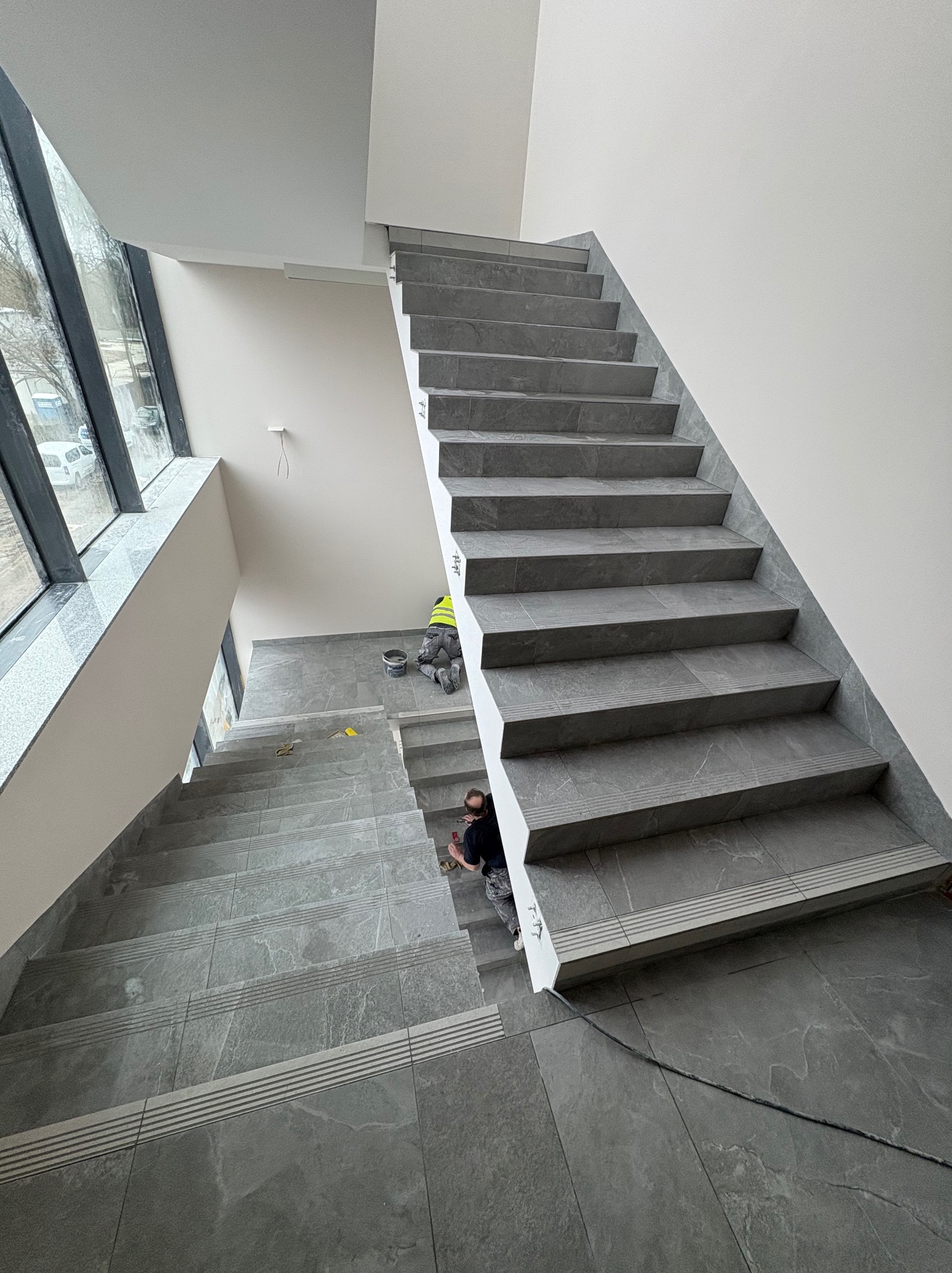 Wykończenie klatki schodowej z szarymi płytkami imitującymi kamień. Widoczni pracownicy podczas fugowania. Duże okno doświetla przestrzeń. Minimalistyczny styl.