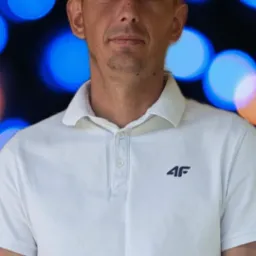 Portret mężczyzny w białej koszulce polo z logo 4F na tle rozmytych świateł, ujęcie od pasa w górę, z lekkim uśmiechem i krótkimi włosami.