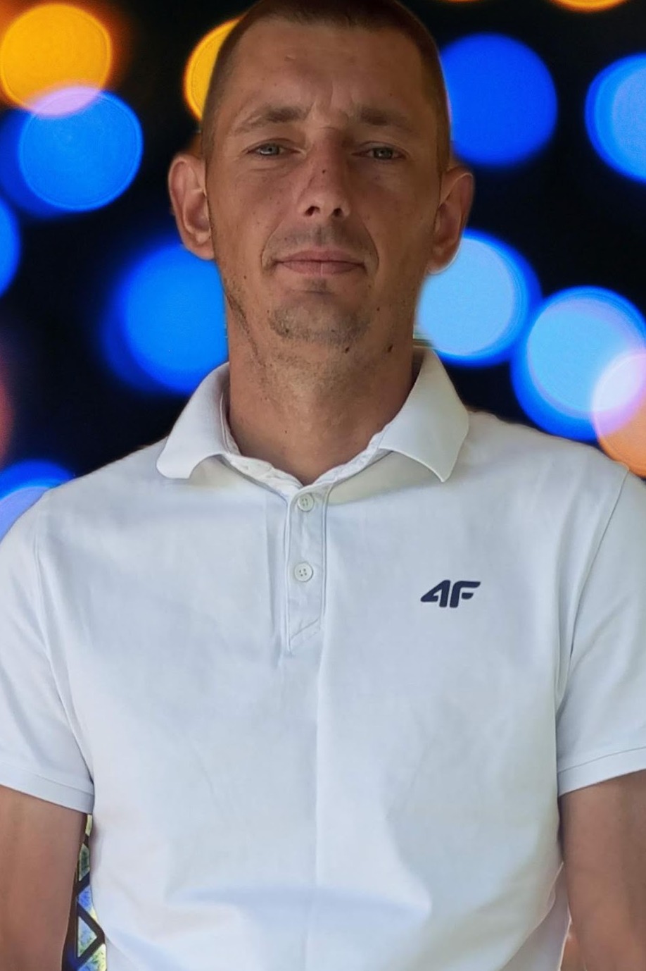 Portret mężczyzny w białej koszulce polo z logo 4F na tle rozmytych świateł, ujęcie od pasa w górę, z lekkim uśmiechem i krótkimi włosami.