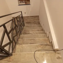 Schody wyłożone płytkami imitującymi marmur, z metalową balustradą o geometrycznym wzorze. Widoczne gniazdka elektryczne i przewody, sugerujące etap wykończeniowy.