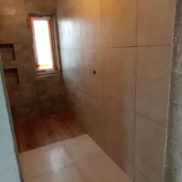 Wykończona łazienka z beżowymi płytkami na ścianach i podłodze. Widoczne okno z pomarańczowym zabezpieczeniem i fragment drewnianej podłogi. Minimalistyczny design.