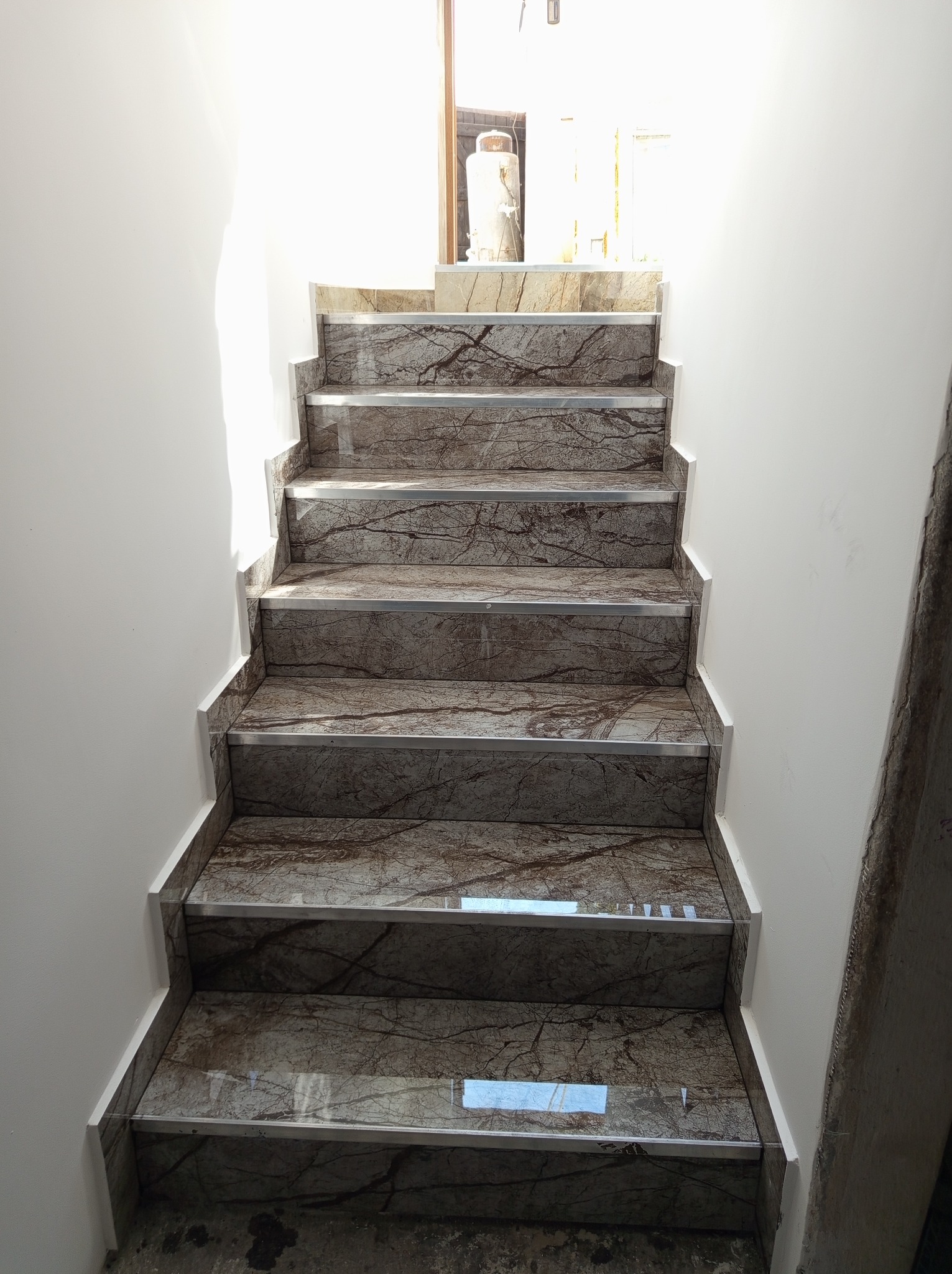 Schody wykończone płytkami imitującymi marmur, z metalowymi listwami, prowadzące do góry. Ściany pomalowane na biało. Widok z dołu.