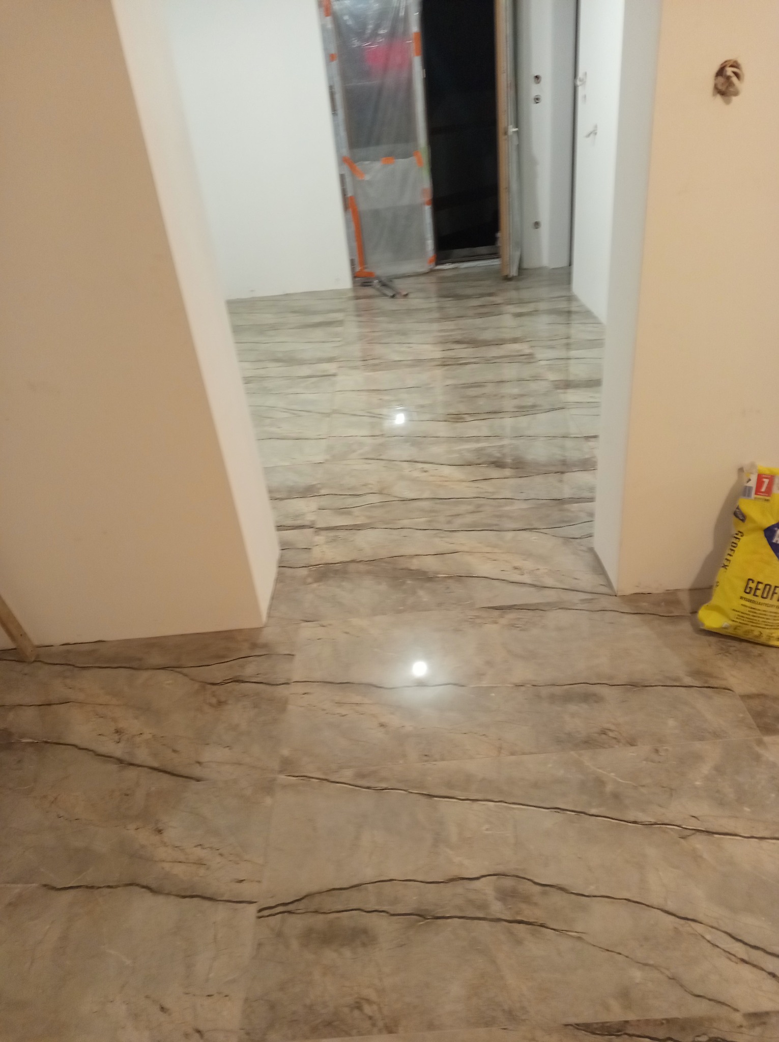 Ułożona podłoga z płytek imitujących marmur, widok na przejście do innego pomieszczenia. W rogu żółty worek z zaprawą klejową do płytek.