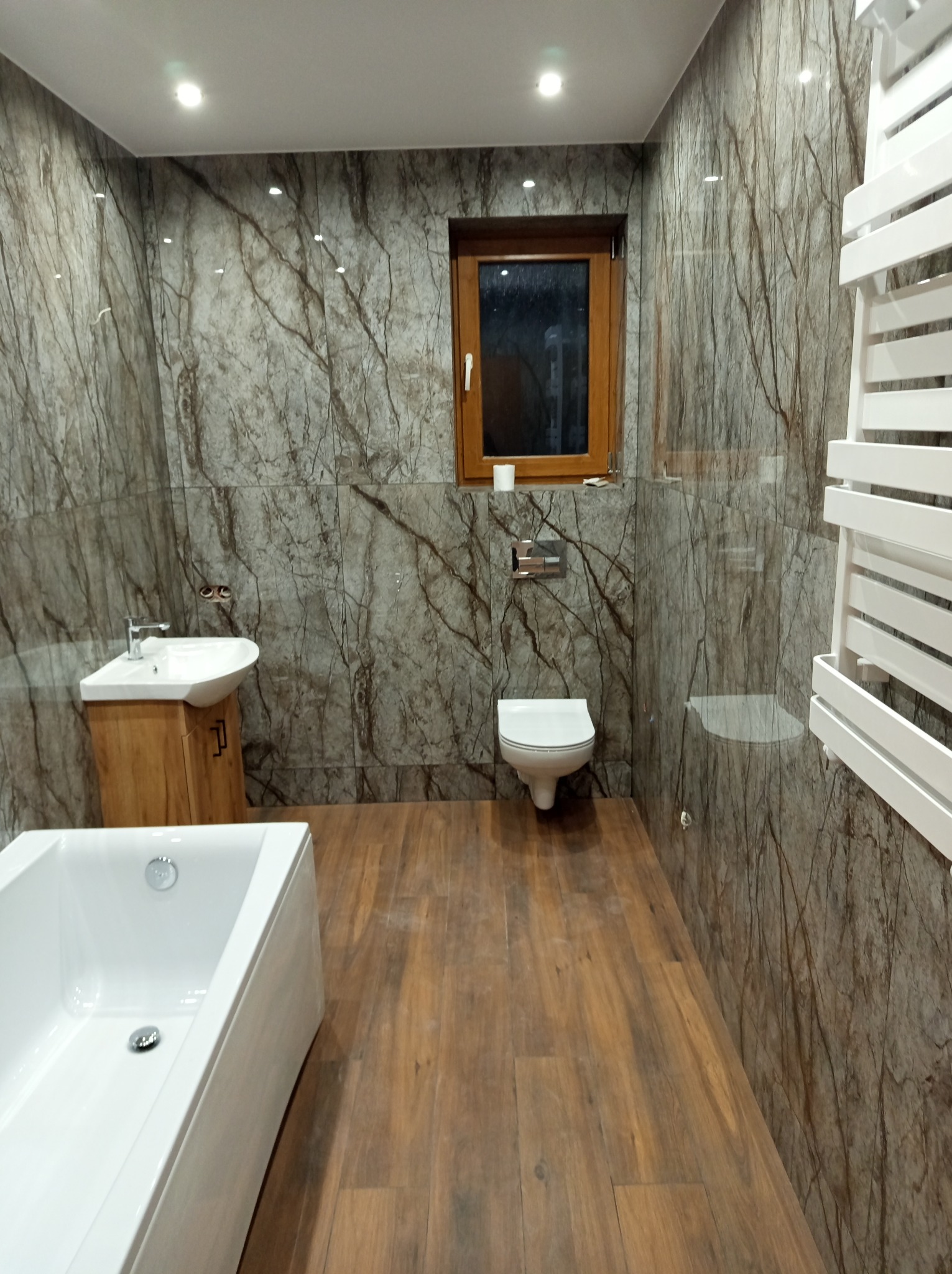 Wykończona łazienka z marmurowymi płytkami, drewnianą podłogą i elementami w kolorze drewna. Wnętrze oświetlone punktowo, z białą armaturą i grzejnikiem drabinkowym.