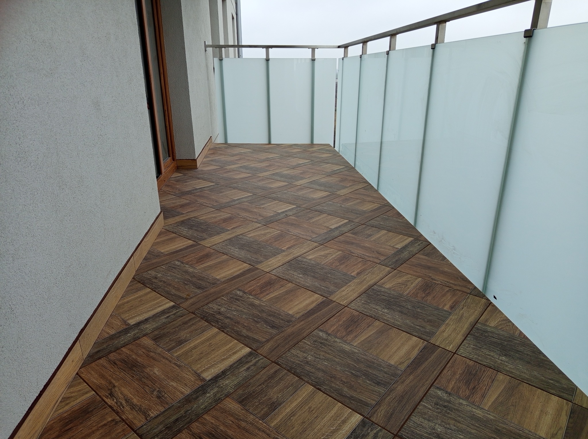 Balkon z drewnopodobną, kwadratową terakotą ułożoną w karo, szklane panele balustrady i szara ściana. Widok z góry.