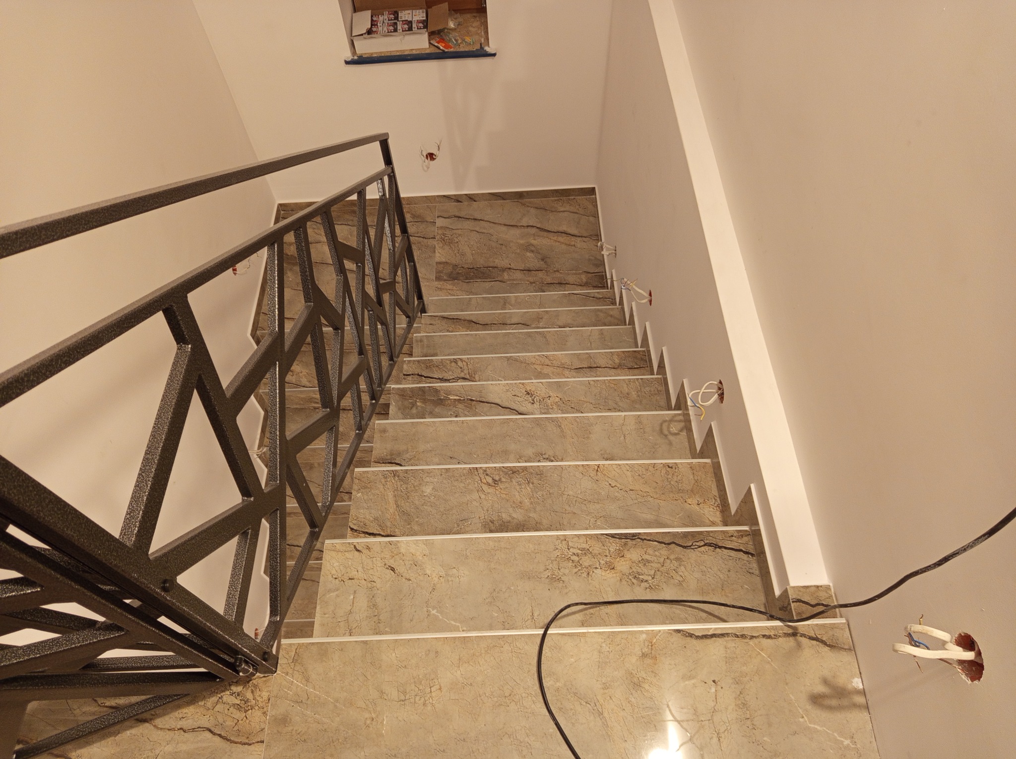 Schody wyłożone płytkami imitującymi marmur, z metalową balustradą o geometrycznym wzorze. Widoczne gniazdka elektryczne i przewody, sugerujące etap wykończeniowy.
