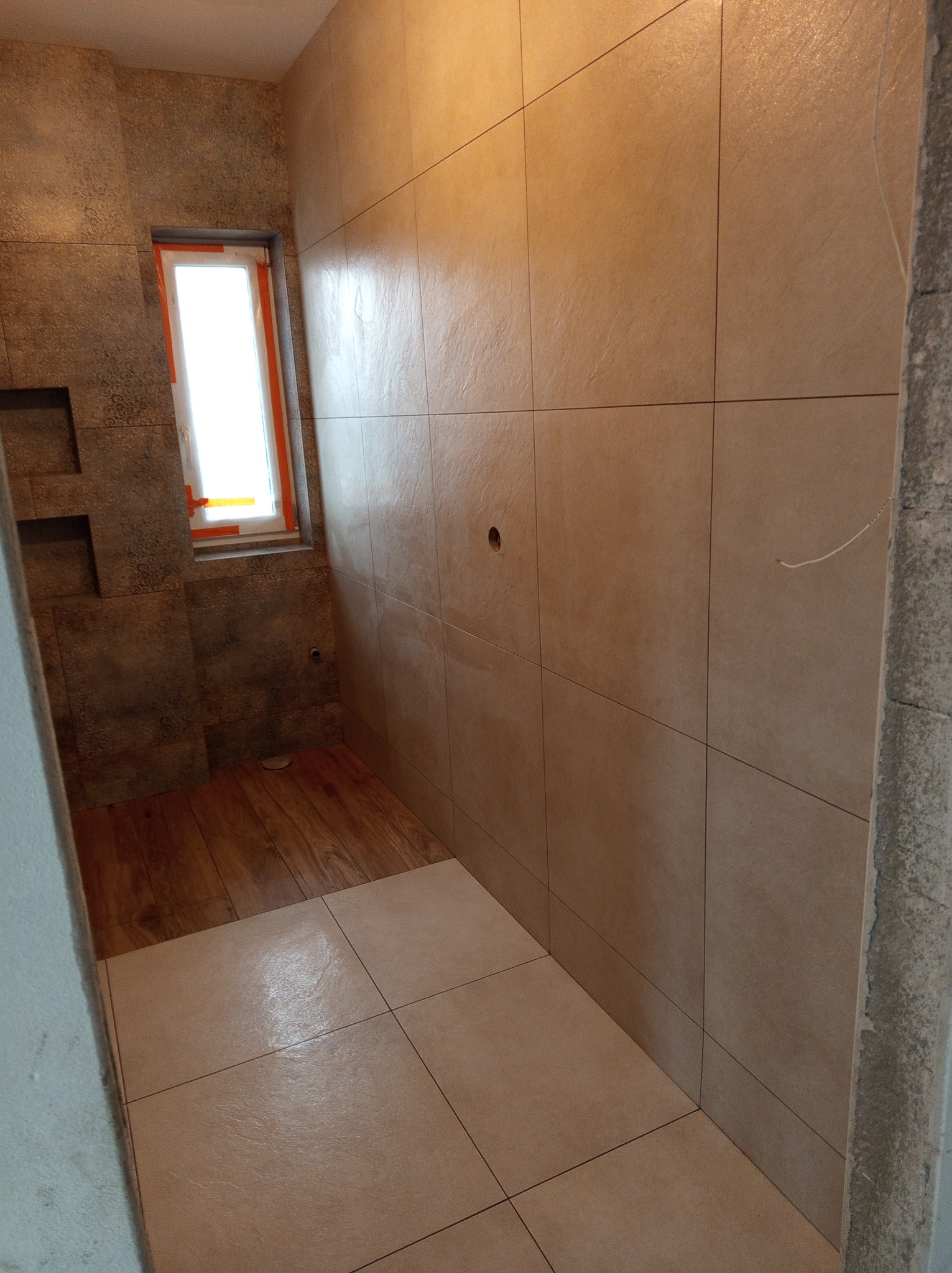 Wykończona łazienka z beżowymi płytkami na ścianach i podłodze. Widoczne okno z pomarańczowym zabezpieczeniem i fragment drewnianej podłogi. Minimalistyczny design.