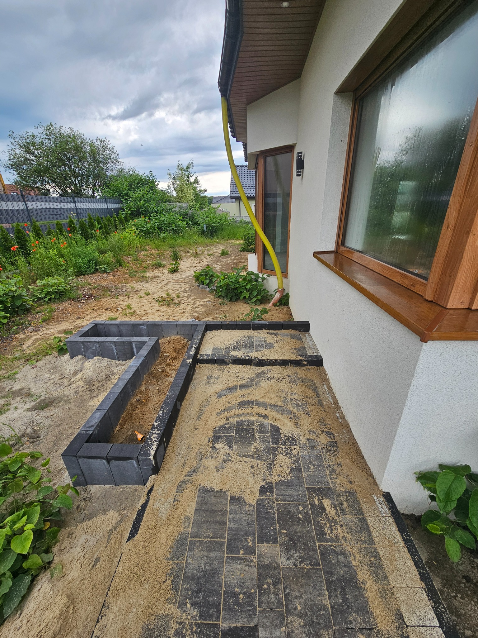 Częściowo ułożona, szara kostka brukowa przy białym budynku z drewnianą ramą okienną. Widoczne przygotowane obrzeża i piasek. W tle ogród z ogrodzeniem.