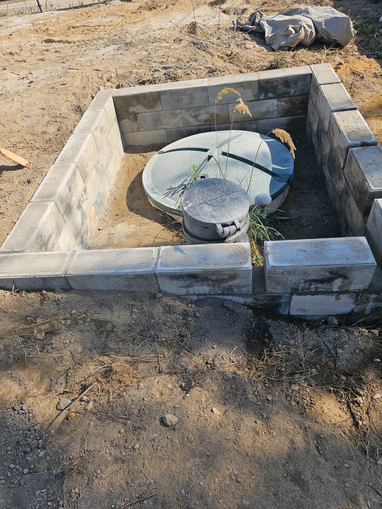 Szambo z betonowych bloczków w ziemi. Okrągła, zielona pokrywa szamba. Na pokrywie czarna, plastikowa nakładka. Dookoła piasek i trawa.