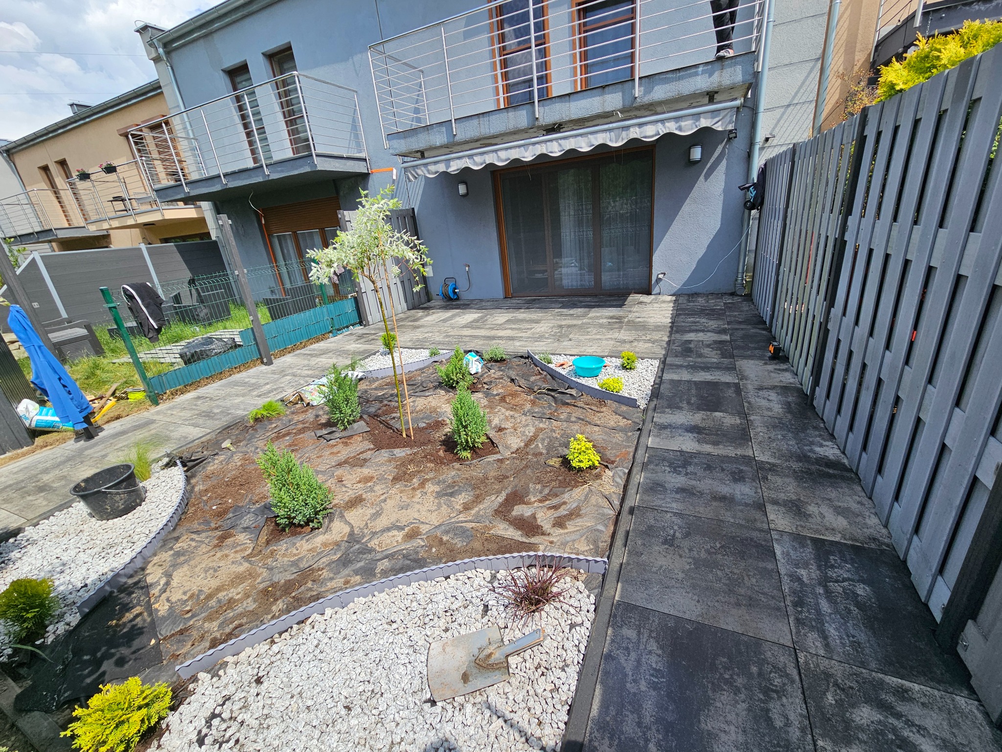 Nowoczesny ogród w trakcie realizacji: czarna kostka brukowa, białe kamienie, sadzonki roślin, szara palisada i metalowy płot na tle domu z balkonem.