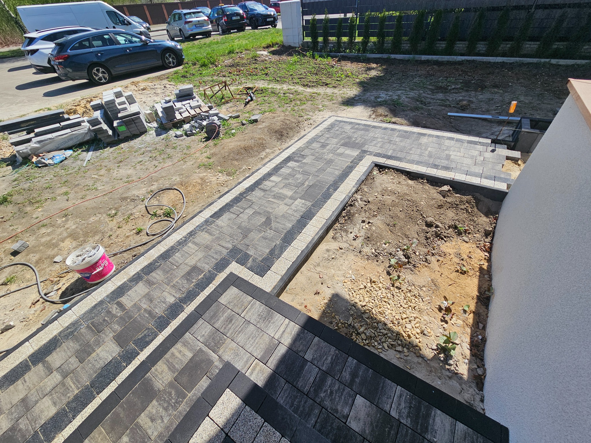 Fragment układanej ścieżki z kostki brukowej w odcieniach szarości, z widocznymi narzędziami i materiałami. Częściowo ukończony projekt na zewnątrz budynku.