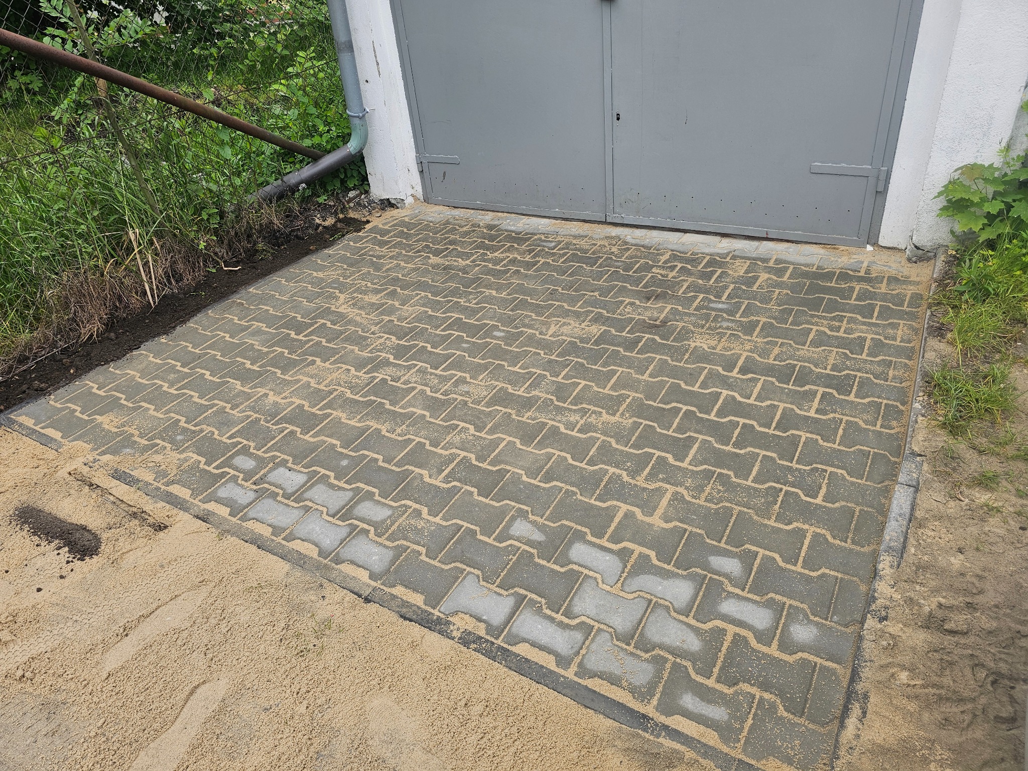 Ułożona kostka brukowa przed garażem, zasypana piaskiem. Szara brama garażowa w tle. Widoczne obrzeża i fragment roślinności.