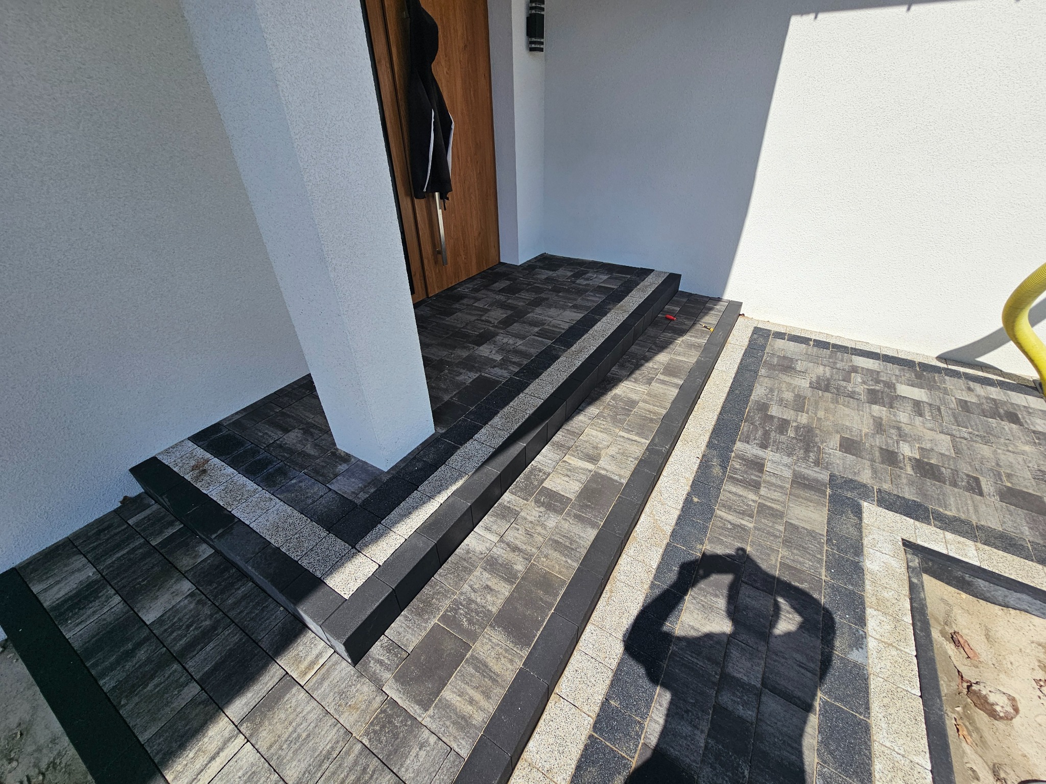 Schody i chodnik z kostki brukowej w odcieniach szarości, z ciemnymi obrzeżami, prowadzące do drzwi wejściowych. Widoczny cień osoby fotografującej.