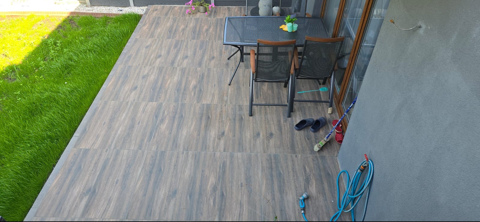 Wykończony taras z brązowych płytek imitujących drewno, zestaw mebli ogrodowych, trawnik. Widok z góry ukazuje detale i estetykę wykonania.