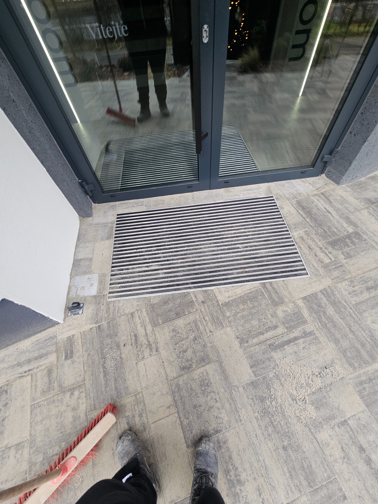 Szare kostki brukowe przed wejściem do budynku z wycieraczką. Widoczna zamiatająca szczotka i fragment buta. Perspektywa z góry.