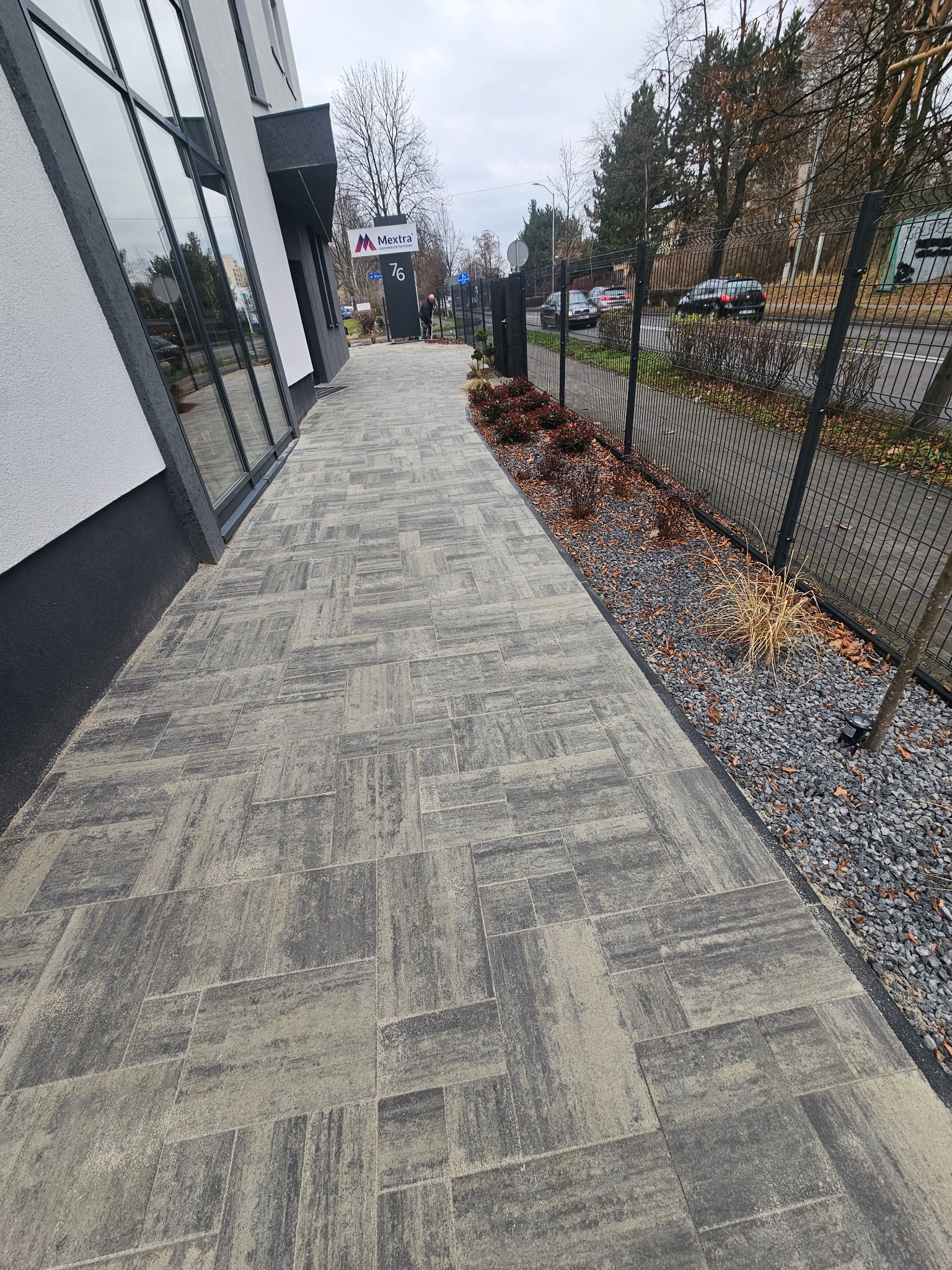 Nowoczesna ścieżka z szarej kostki brukowej o nieregularnym wzorze, prowadząca wzdłuż budynku z dużymi oknami i ogrodzeniem panelowym. Widoczne nasadzenia roślin.