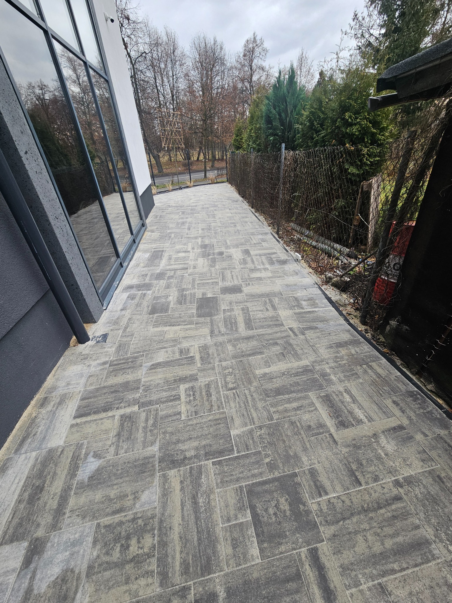 Ułożona kostka brukowa w odcieniach szarości tworzy ścieżkę obok nowoczesnego budynku z dużymi oknami i siatkowym ogrodzeniem w tle.