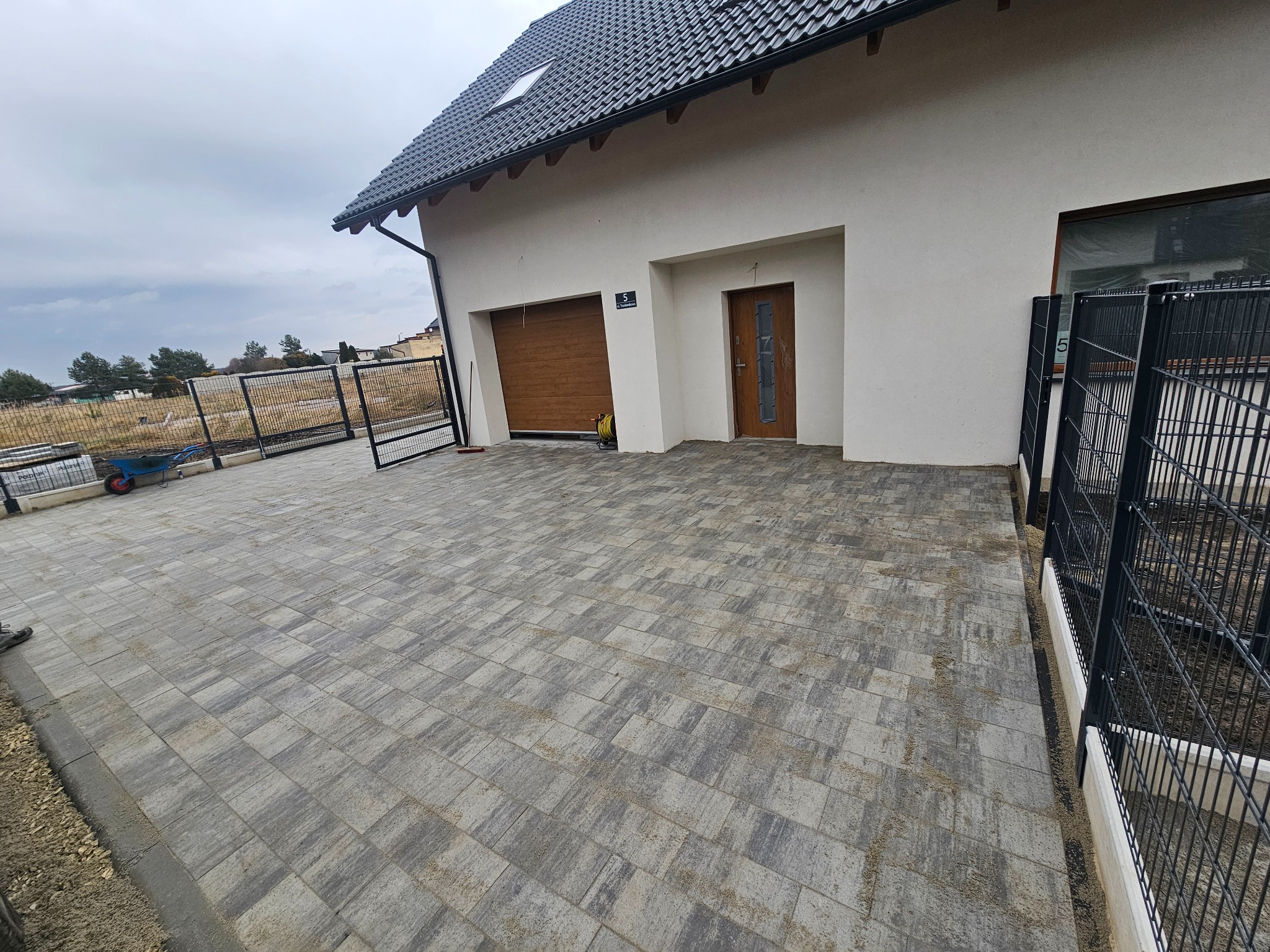 Ułożona kostka brukowa w odcieniach szarości przed nowoczesnym domem z brązową bramą garażową i czarnym ogrodzeniem panelowym. Widok z perspektywy.