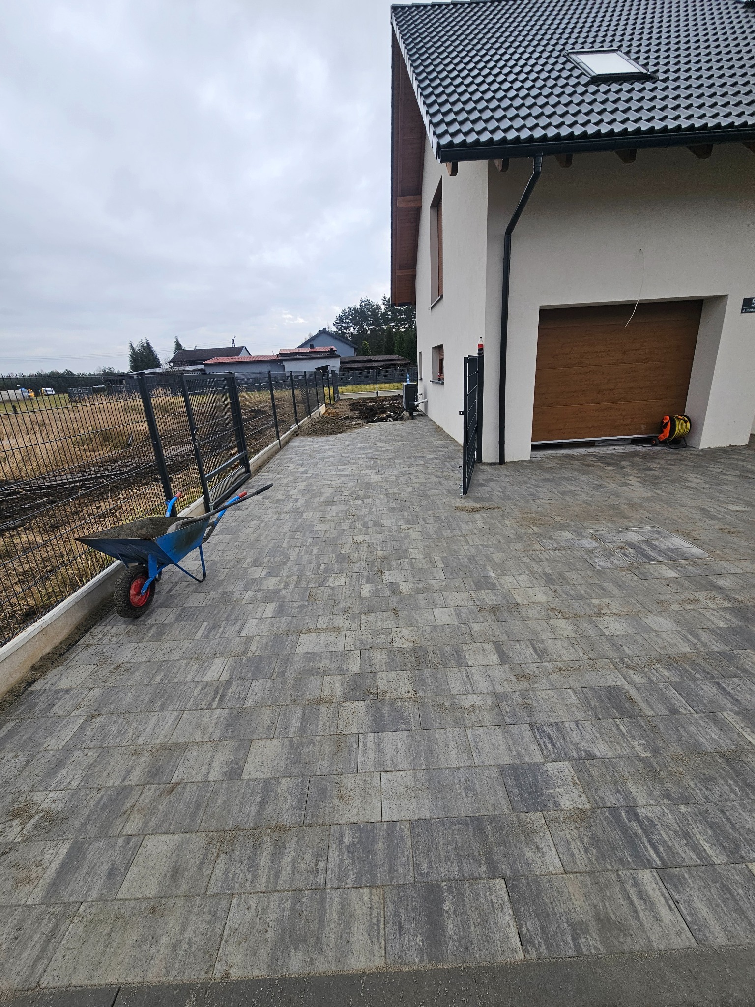 Ułożona kostka brukowa w szarym odcieniu przed domem z brązową bramą garażową. W tle widoczne ogrodzenie panelowe i częściowo przygotowany teren.