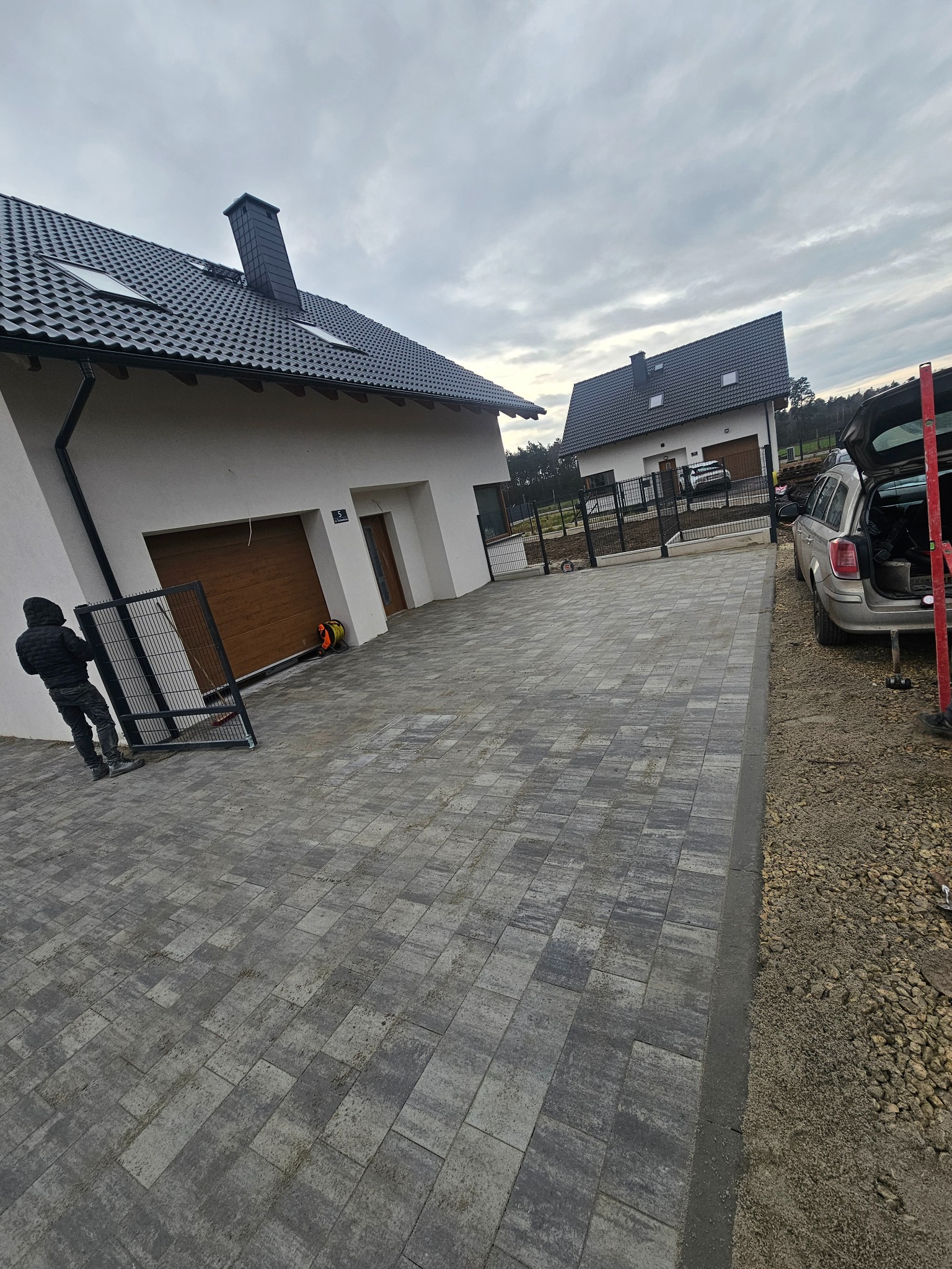 Ułożona kostka brukowa na podjeździe do domu, widoczny montaż ogrodzenia panelowego, samochód z otwartym bagażnikiem i narzędzia. Dwa domy w tle.