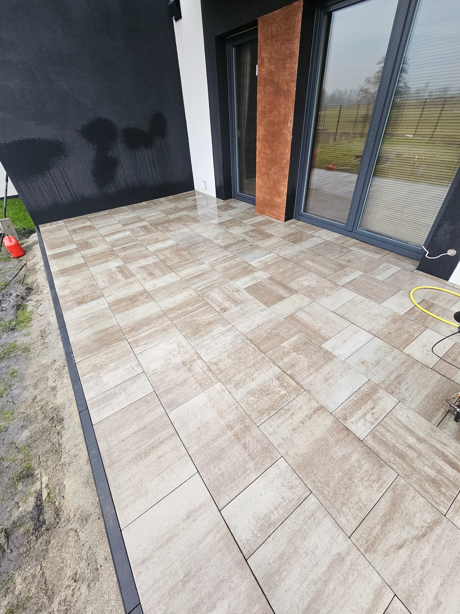 Nowoczesny taras z jasnej kostki brukowej ułożonej wzorem prostokątnym, z ciemną obwódką. Widoczne okna tarasowe i fragment czarnej ściany budynku.