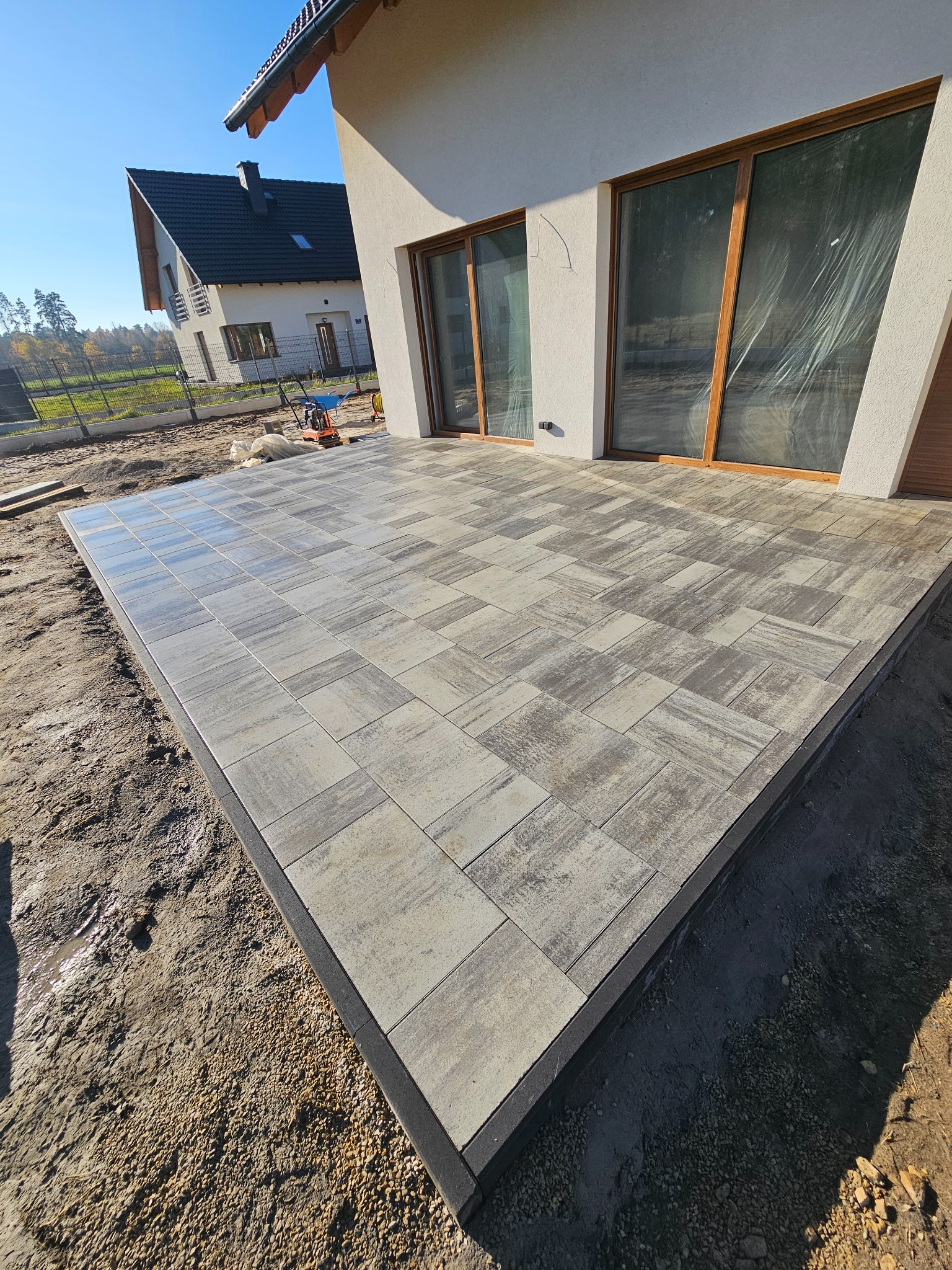 Nowoczesny taras z kostki brukowej w odcieniach szarości, z czarnym obrzeżem, przylegający do jasnego budynku z dużymi oknami. W tle widoczny drugi budynek i maszyna budowlana.