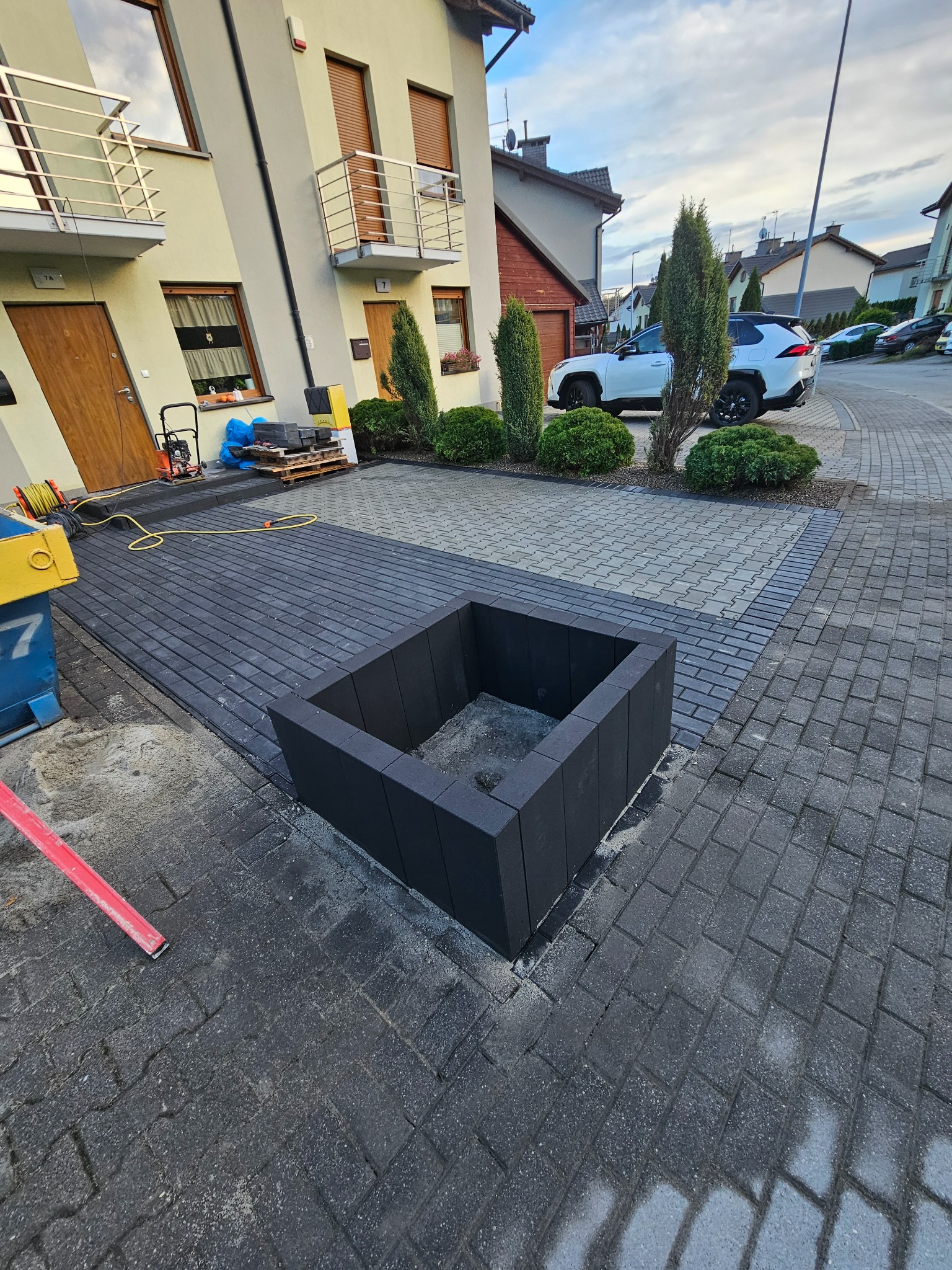 Ułożona, ciemnoszara kostka brukowa z czarną donicą w centrum, na tle domu z beżową elewacją i zieleni. Widoczny fragment narzędzi brukarskich.