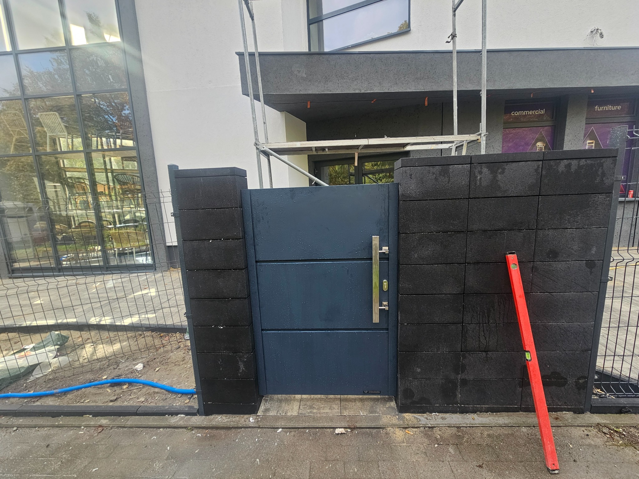 Nowoczesna, grafitowa furtka z poziomym wzorem, wkomponowana w ogrodzenie z czarnych, betonowych bloczków. Czerwona poziomica oparta o słupek. Widoczne rusztowanie.