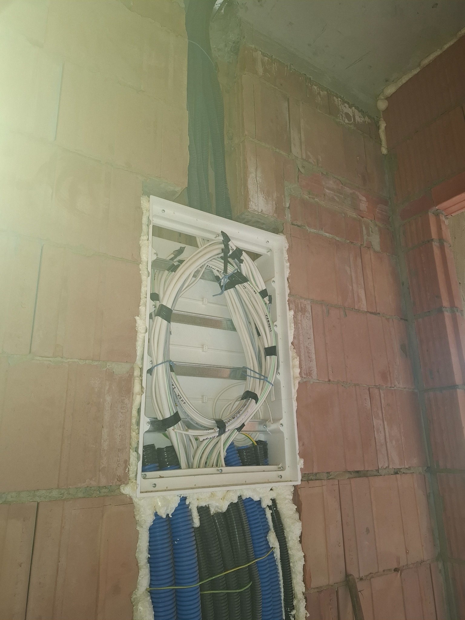 Puszka elektryczna w ścianie z kablami, zabezpieczona pianką montażową. Widoczne peszle z kablami. Surowy mur z cegły. Etap prac instalacyjnych.