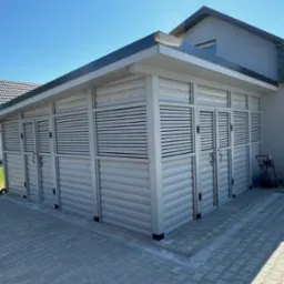 Biała, drewniana altana z żaluzjowymi drzwiami na kostce brukowej, przylegająca do budynku. Widoczne detale wykończenia i elementy otoczenia.