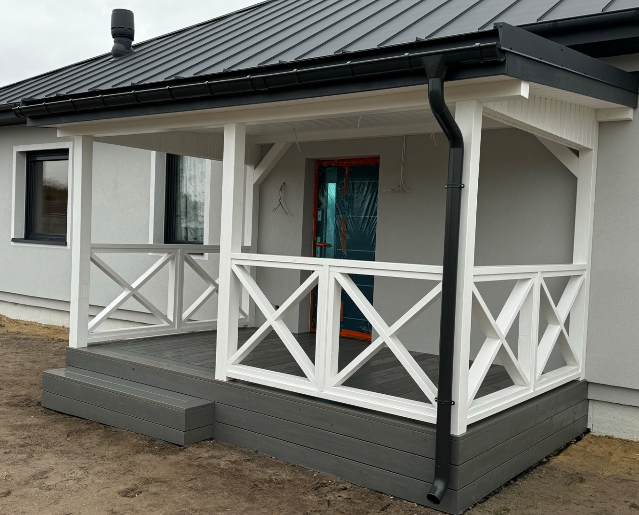 Zadaszony taras z drewnianą balustradą w kształcie krzyży, szare deski podłogowe i białe słupy. Widoczne schody wejściowe i fragment elewacji domu.