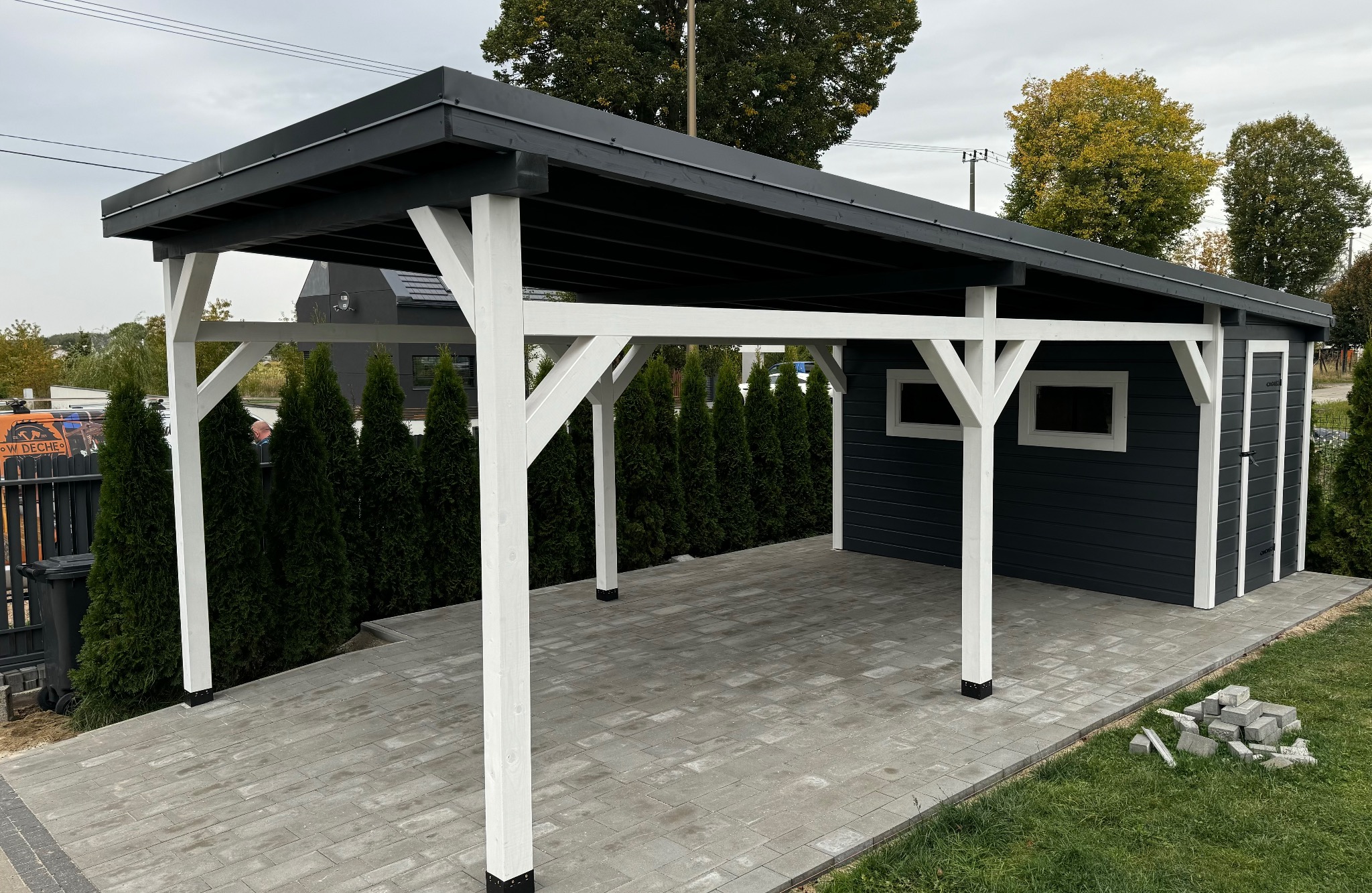 Nowoczesna, grafitowo-biała konstrukcja z drewna z zadaszeniem, prawdopodobnie wiata garażowa, na tle kostki brukowej i zielonych tuj.