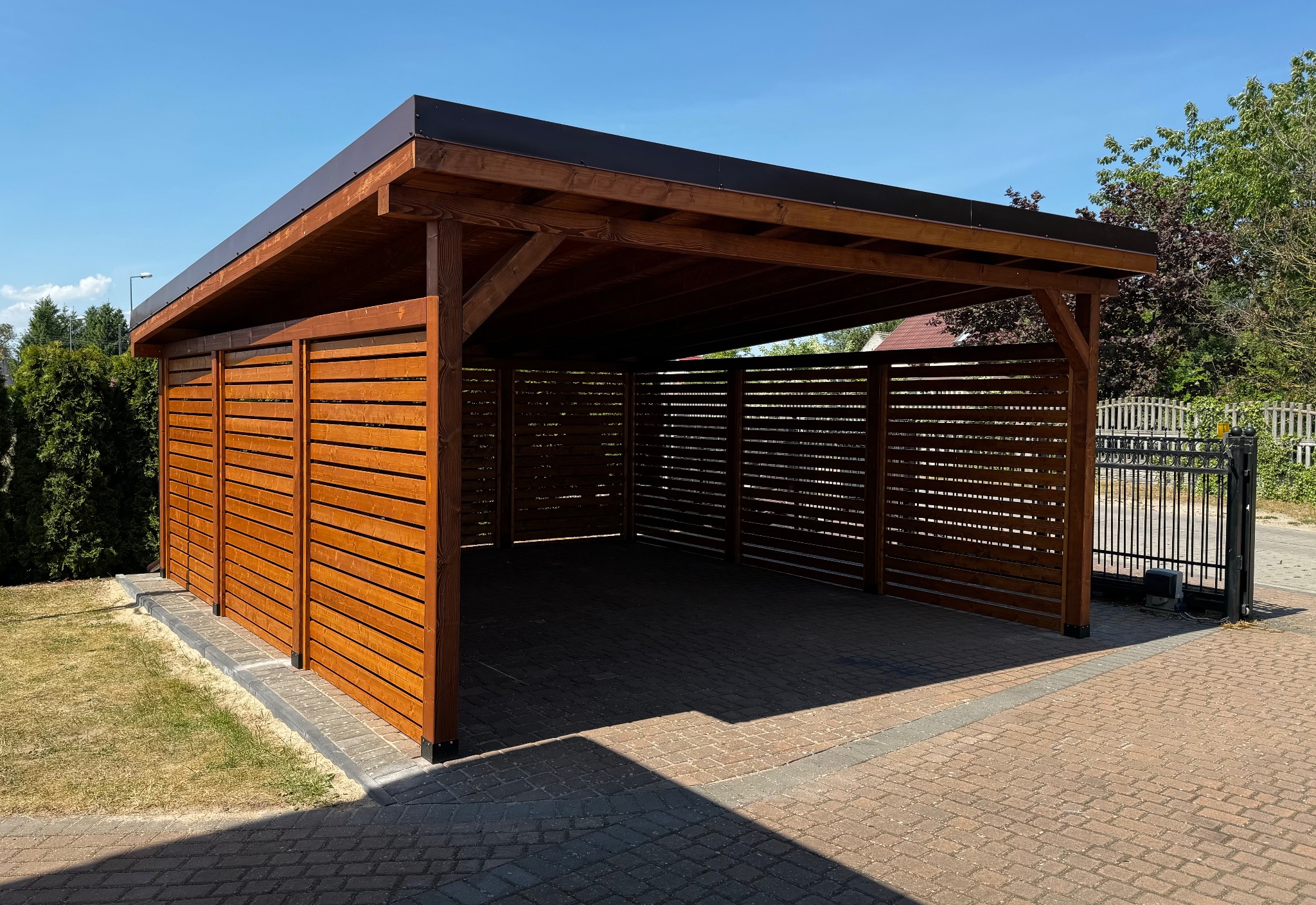 Zadaszenie drewniane z brązowym dachem, osłonięte z trzech stron poziomymi listwami, na tle kostki brukowej i zieleni. Widok z zewnątrz, w słoneczny dzień.
