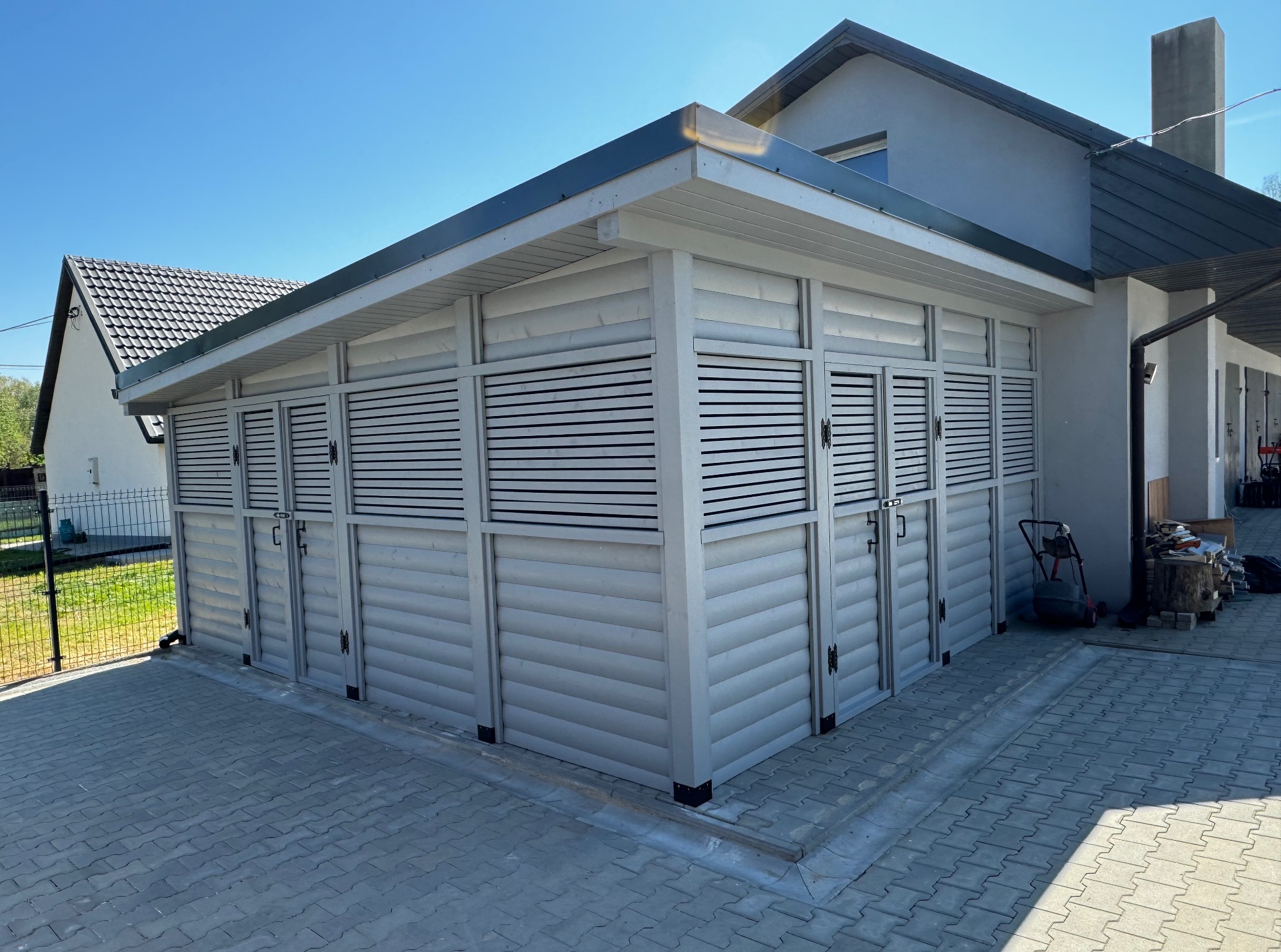 Biała, drewniana altana z żaluzjowymi drzwiami na kostce brukowej, przylegająca do budynku. Widoczne detale wykończenia i elementy otoczenia.