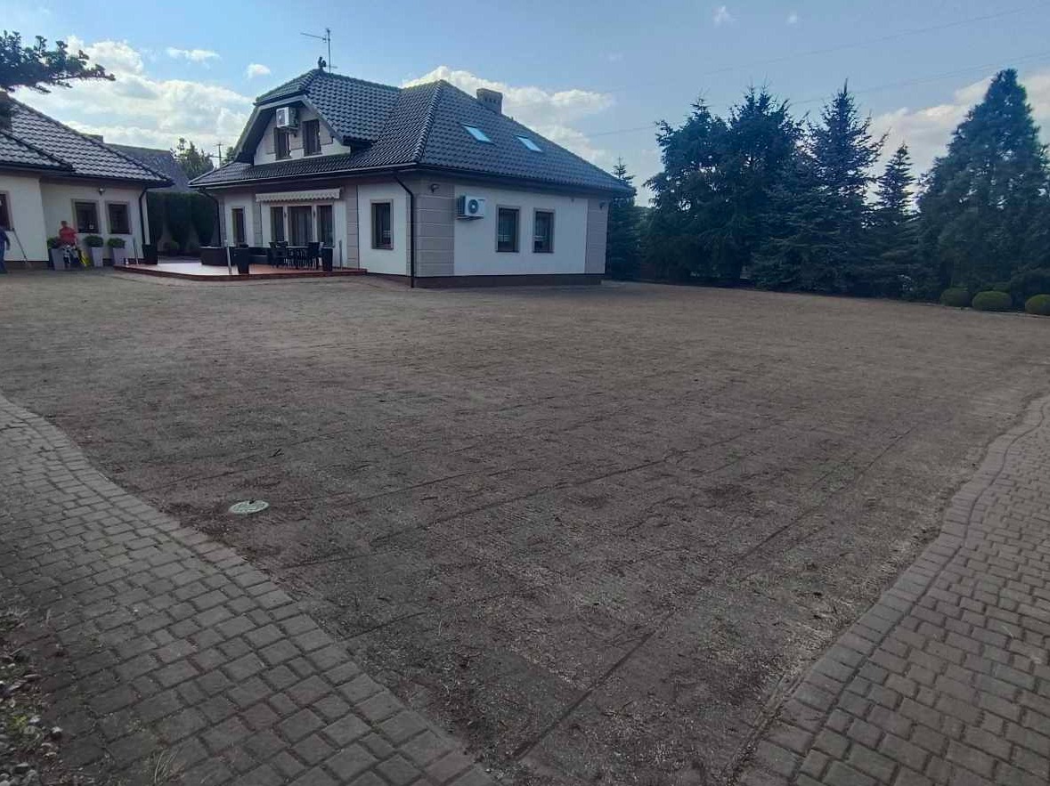 Przygotowany teren pod trawnik przed domem z kostką brukową i zielenią w tle. Widoczne ślady po maszynach, grunt wyrównany i gotowy do siewu lub położenia trawy z rolki.