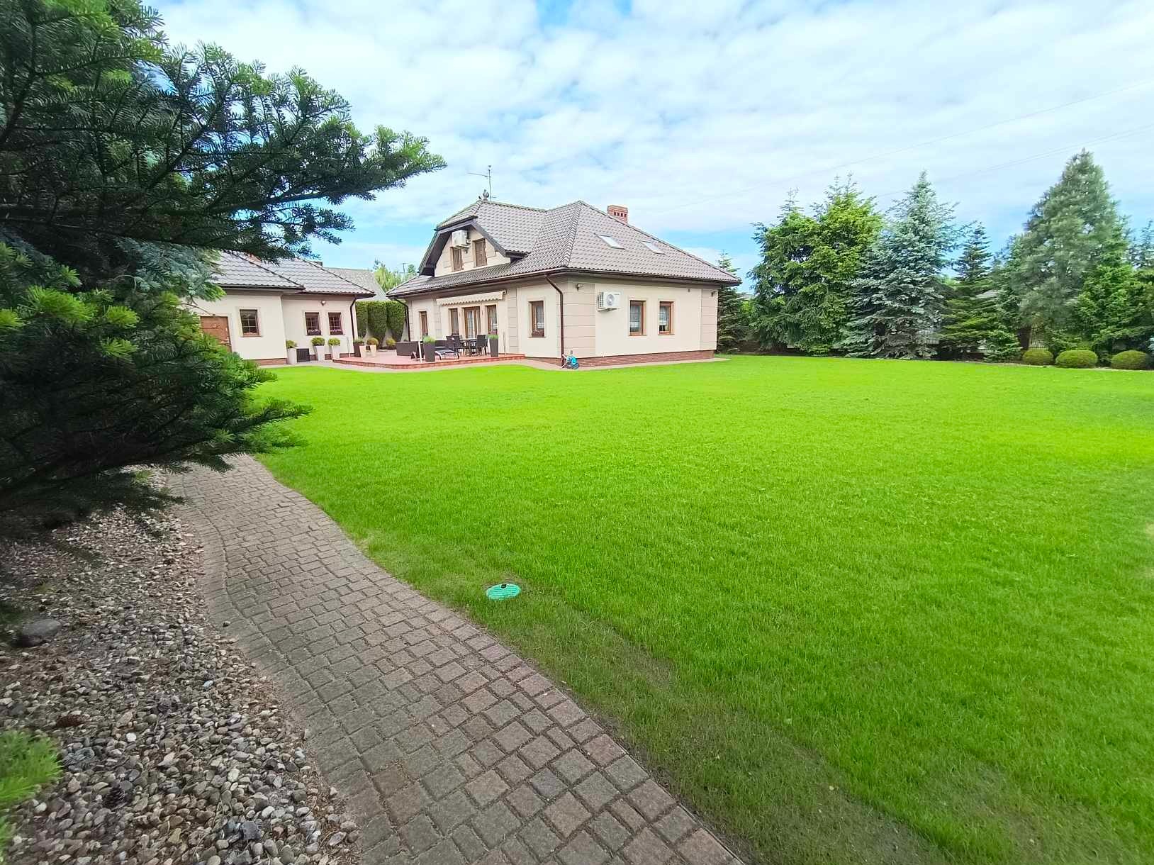 Zadbany trawnik z równo skoszoną trawą, widok z kamiennej ścieżki. W tle dom z ogrodem w Rokietnicy. Widoczne drzewa iglaste.