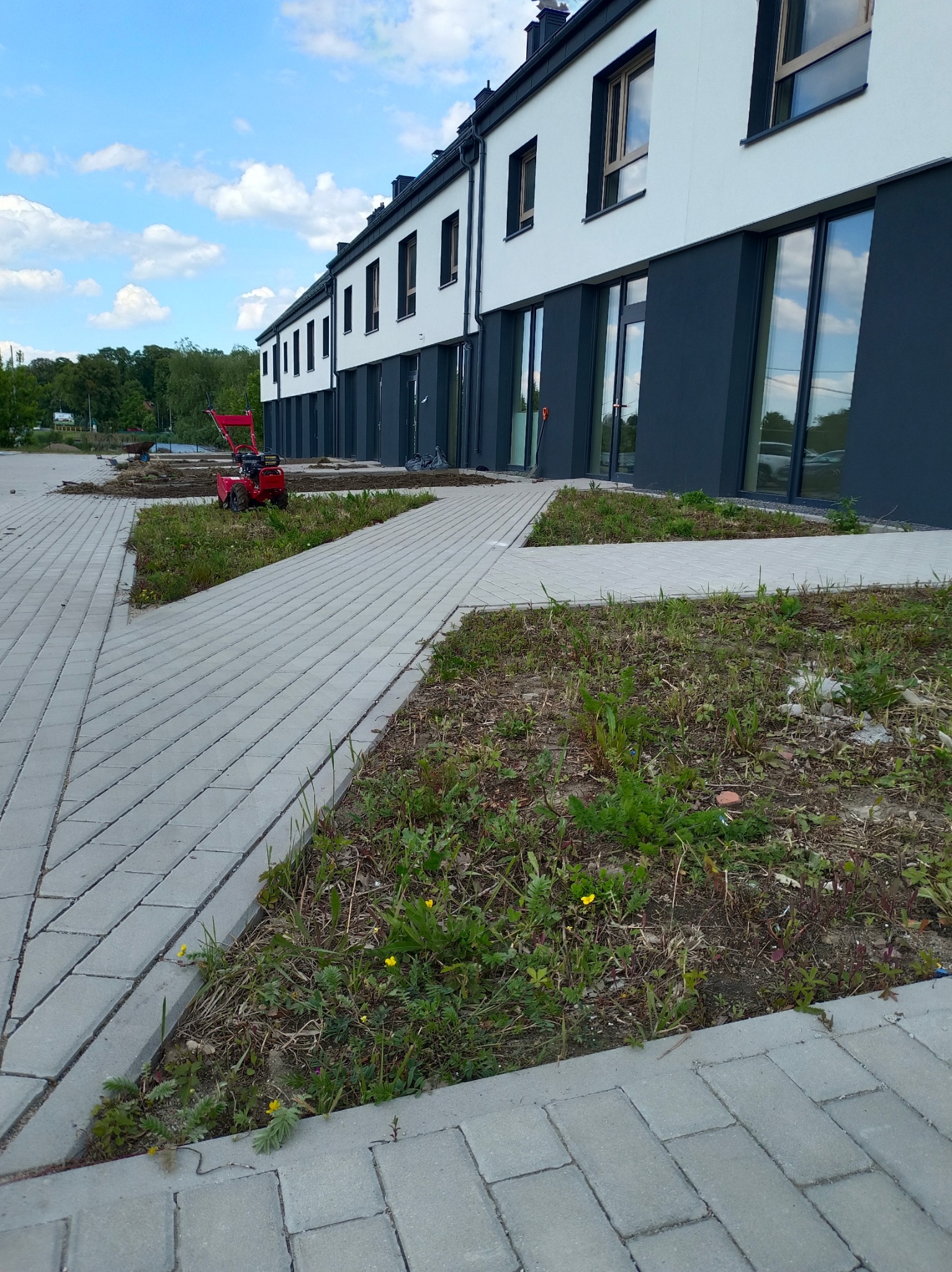 Przygotowanie terenu pod ogród przy nowoczesnym budynku. Czerwony glebogryzarek stoi na ziemi, obok ułożona kostka brukowa, w tle drzewa i błękitne niebo.