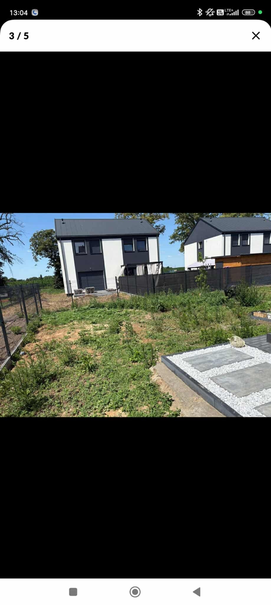 Działka przed domem z widocznymi chwastami i fragmentem ścieżki z płyt betonowych i białego żwiru. W tle dwa nowoczesne domy w biało-czarnej kolorystyce.