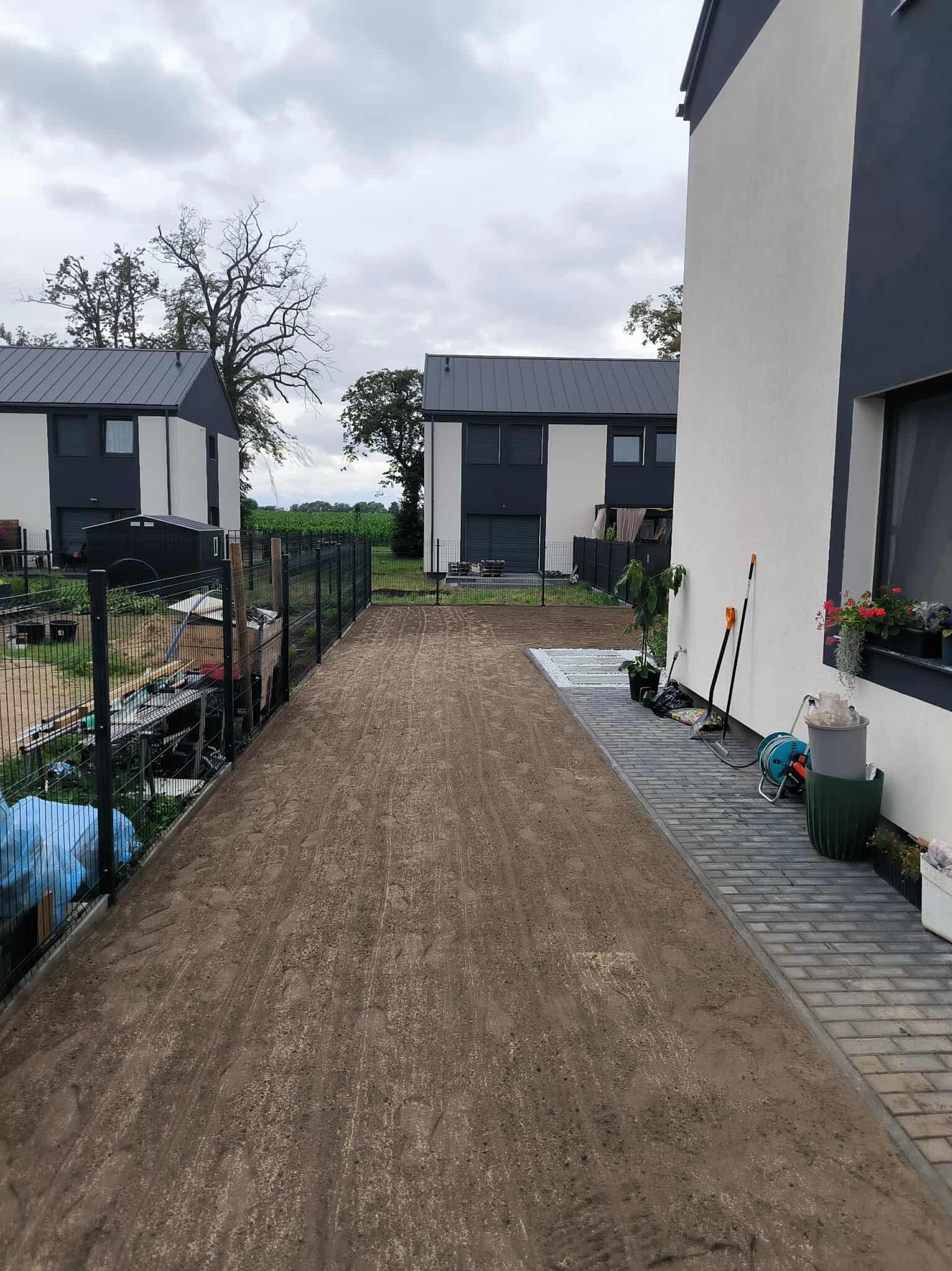 Przygotowany teren pod trawnik między dwoma nowoczesnymi domami. Widoczne narzędzia ogrodnicze i donice. Trawa jeszcze niezasiana, ziemia wyrównana.